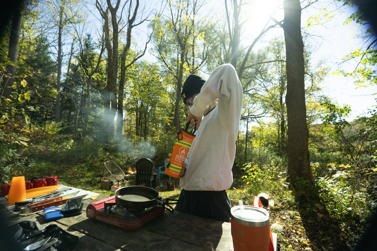 Person holding a FireSafe fire blanket near a camp stove with a flaming pan in a forest