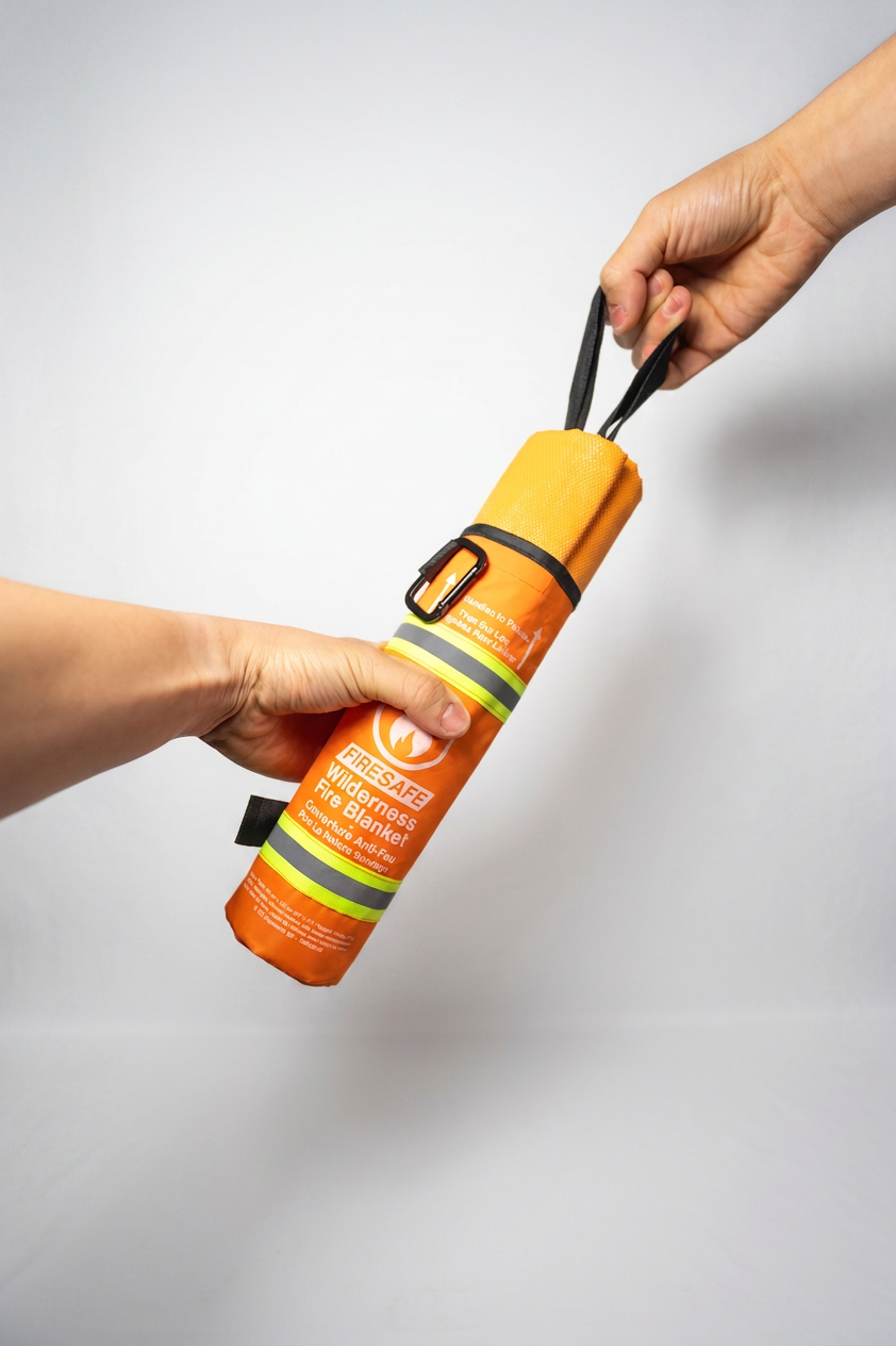 Two hands holding the FireSafe Wilderness Fire Blanket showing the orange cylindrical housing and pull handle on clean white backdrop
