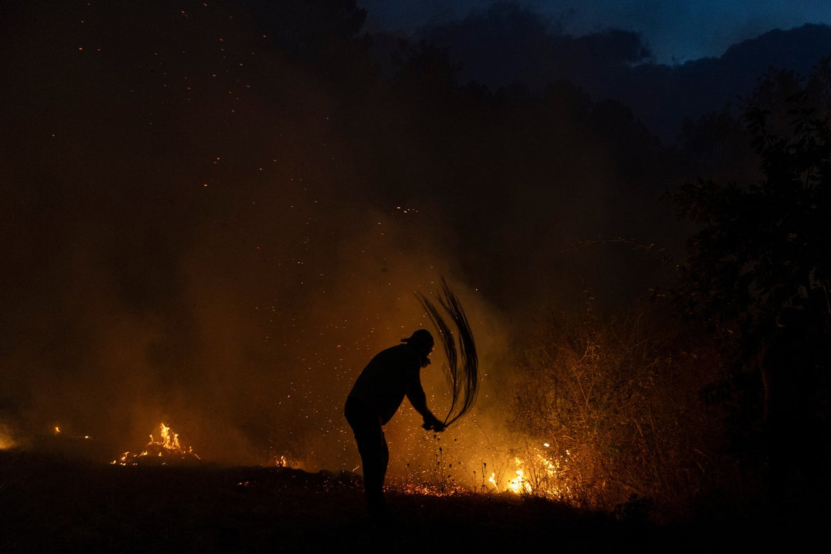Person swinging a fire-beater at ground flames amid sparks and smoke at twilight